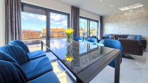 A modern living room with a glass-top coffee table, blue sofas, and a brown sofa with patterned cushions. - Penthouse Rosa (Radici)