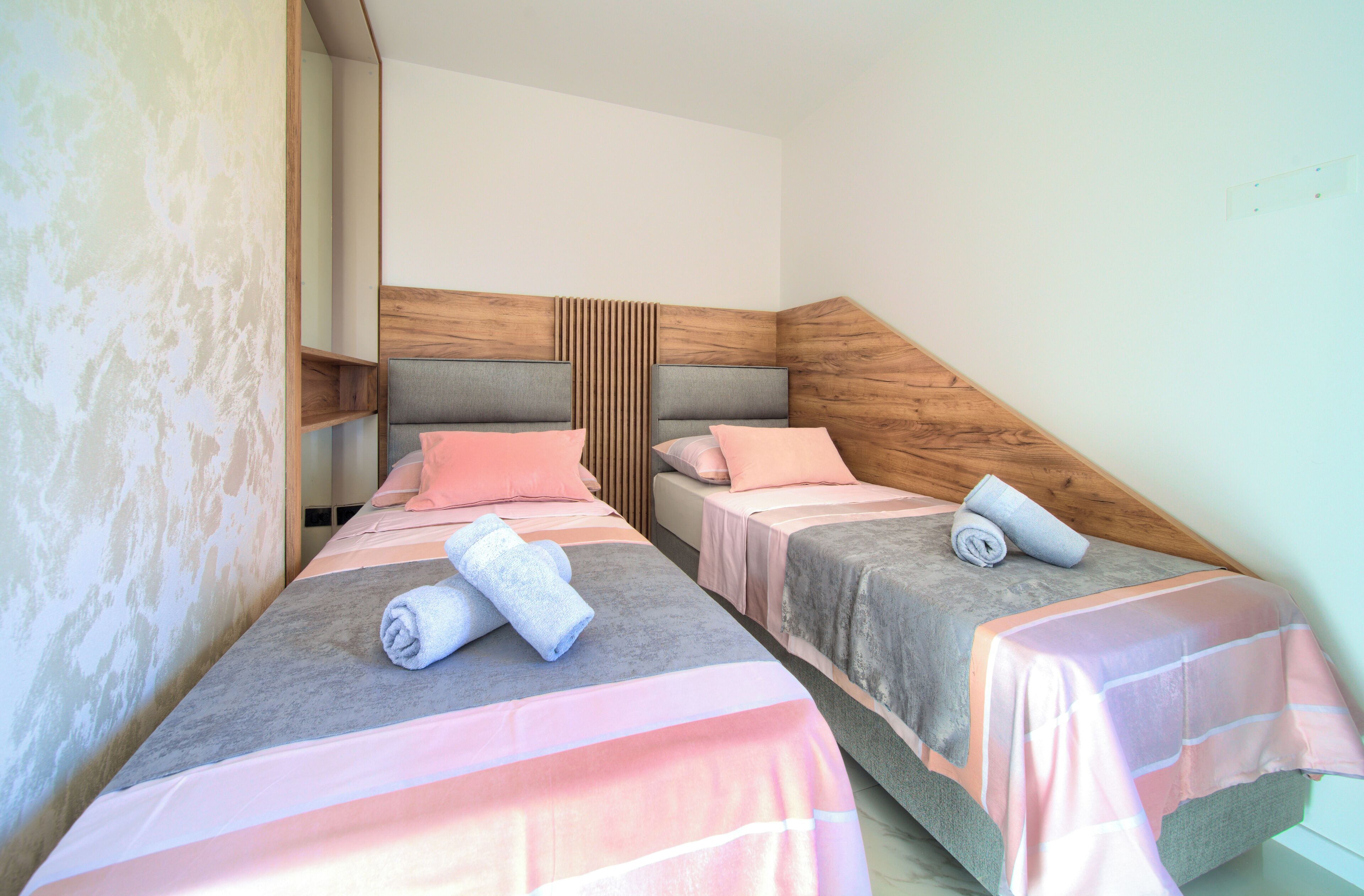 A hotel room with two single beds, each with a wooden headboard, a grey and pink striped bedspread, and rolled towels on the beds.