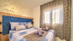 A bedroom with a blue-striped bedspread, a wooden headboard, and a window with curtains. - Penthouse Rosa (Radici)