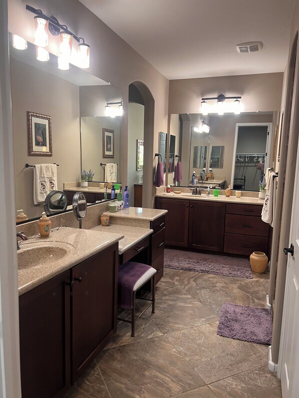 A bathroom with two sinks, a vanity, and a large mirror. There are multiple toiletries on the counter, and a purple rug on the floor.
