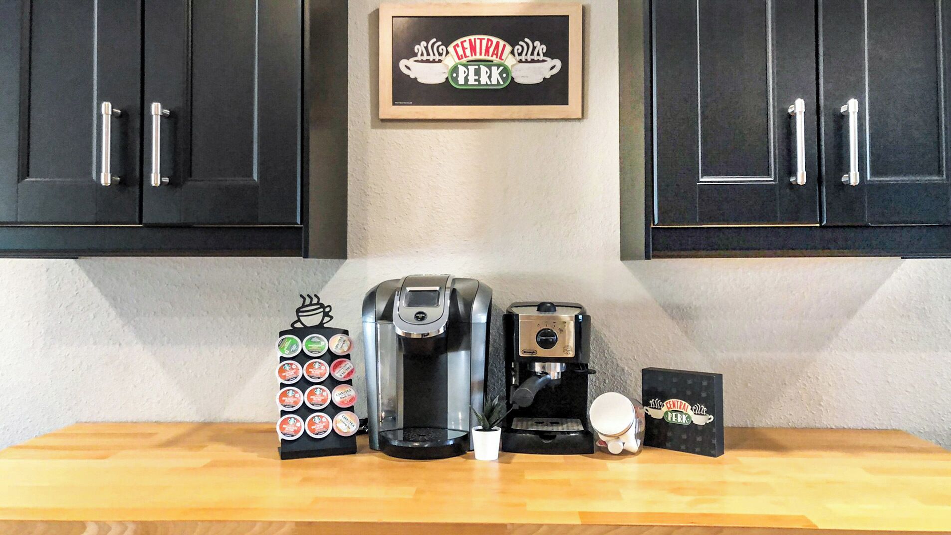 Un comptoir de cuisine avec une machine à café, un grille-pain, une tasse, un support à dosettes de café et un cadre décoratif portant l’inscription « Central Perk ».