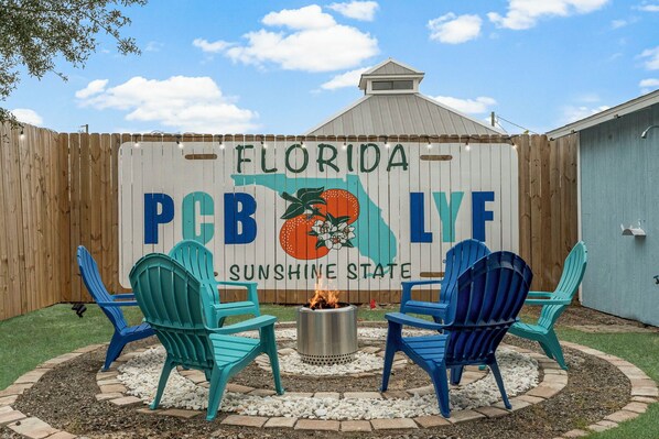 Takapihalla on puinen aita, kaksi Adirondack-tuolia, nuotiopaikka ja kyltti, jossa lukee: ”Florida PCB LYF, auringonpaisteen osavaltio”.