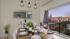 Un coin repas avec une table en bois dressée pour quatre personnes, un vase de fleurs et une bouteille de vin. Un grand miroir est accroché au-dessus de la table, et un balcon offre une vue sur les bâtiments.