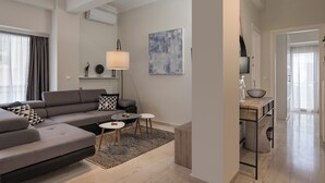Un salon moderne avec un canapé d’angle gris, une petite table basse et une lampe. Un tableau orne le mur, et une fenêtre est agrémentée de rideaux.