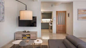 Un salon moderne avec un canapé, une table basse et un téléviseur. Une porte en bois donne accès à une cuisine et à un coin repas.