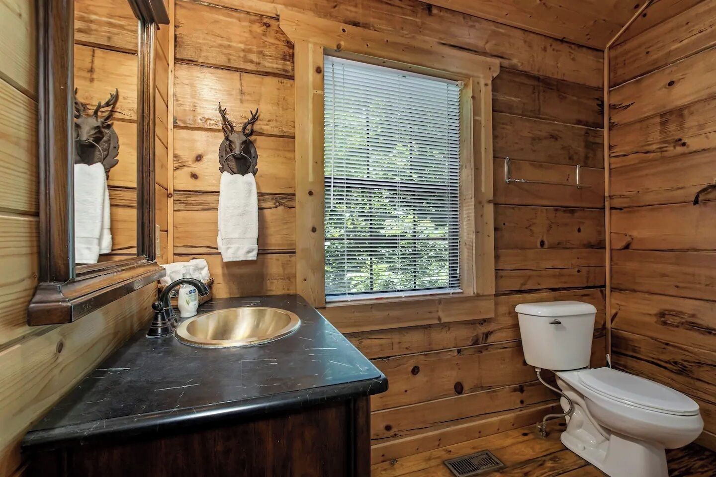 A wooden cabin bathroom with a sink, toilet, and window with blinds.