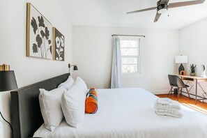 A hotel room with a bed, a desk, a chair, a ceiling fan, and artwork on the wall.