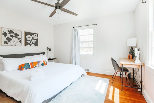 Un dormitorio con cama, escritorio, silla, ventilador de techo y dos obras de arte colgadas en la pared.
