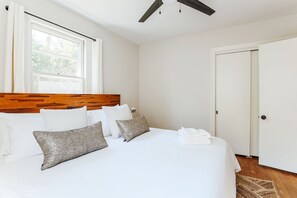 Una cama bien hecha con sábanas blancas y dos almohadas, un cabecero de madera, una ventana con cortinas, un ventilador de techo y una puerta.
