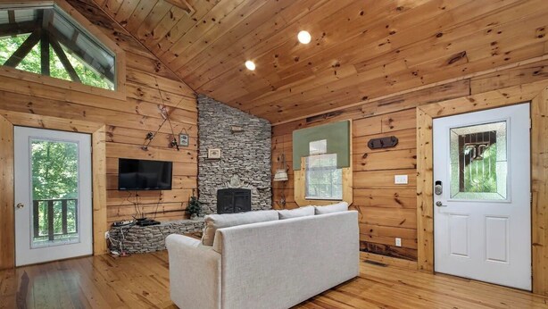Interior acolhedor de uma cabana com paredes de madeira, lareira de pedra, TV de tela plana e um sofá.
