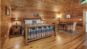 Quarto de cabana de madeira com cama, duas luminárias de cabeceira, uma mesa e uma cadeira.