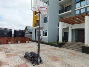 A basketball hoop in an outdoor courtyard with a tiled floor, a bench, and a building with balconies and a pergola. - 2,3 Ensuite Bedrooms Luxury Staycation and Vacation apartments (Kasoa)