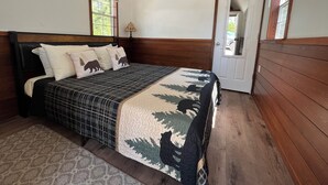 Un dormitorio con cabecera de madera, cama con edredón estampado, dos almohadas con motivos de osos, una mesita de noche, una lámpara y una puerta que da a otra habitación.