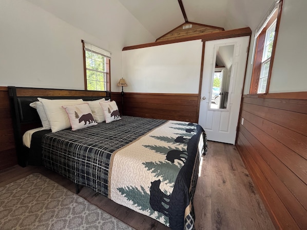 Une chambre à coucher avec une tête de lit en bois, un lit recouvert d’une couvre-lit à motifs, deux oreillers ornés de motifs d’ours, une table de chevet, une lampe et une porte donnant sur une autre pièce.