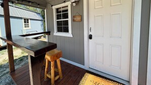 Una terraza de madera con un banco y un taburete pequeño, una puerta blanca con un buzón, y una ventana con un cartel que dice 'Quítese los zapatos'.