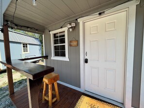Eine Holzterrasse mit einer Bank und einem kleinen Hocker, eine weiße Tür mit einem Briefkasten und ein Fenster mit einem Schild, auf dem 'Schuhe ausziehen' steht.