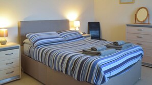 Una cama bien hecha con ropa de cama a rayas, una lámpara de noche, una silla, un aparador con cajones y un cuadro en la pared.