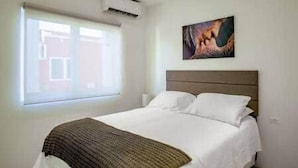 A bedroom with a bed, a window, an air conditioning unit, and a painting on the wall.