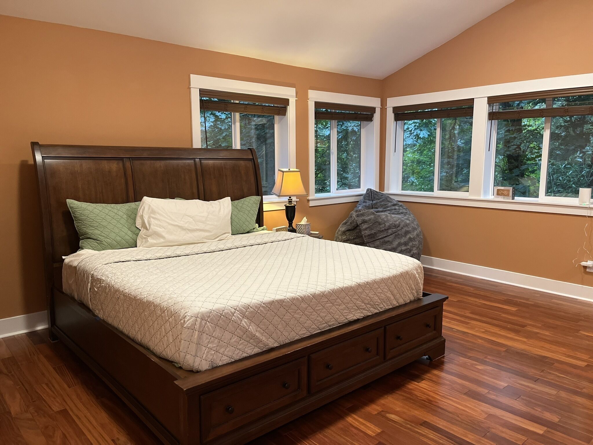 A bedroom with a wooden bed, a nightstand with a lamp, and a view of trees through large windows.