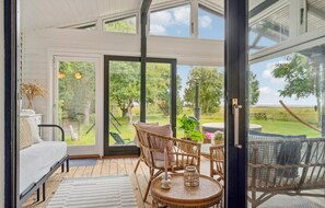 Ruime kamer met een groot schuifglas dat uitkomt op een buitenruimte. Binnenin staat een bed, een rieten stoel en een klein rond tafeltje met een kaars en een glazen pot.