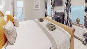 A neatly made bed with a white comforter and a wooden headboard, a bedside table with a lamp, and a view of a fireplace through a window with floral curtains. - 4 bedroom accommodation in Coxhoe, nearby Durham (Coxhoe, nearby Durham)