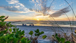 Escena de playa con atardecer, mar tranquilo y gente relajándose en la orilla.