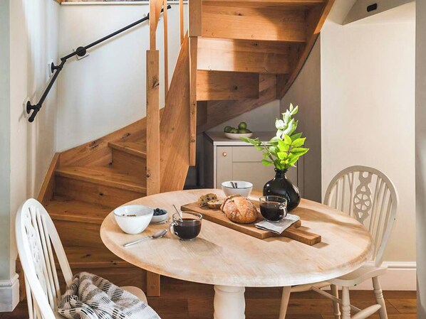 A dining area with a round table, chairs, a vase with flowers, a bowl of cereal, a cup of coffee, and a loaf of bread.
