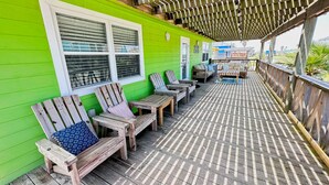 A covered wooden deck with Adirondack chairs and a table, adjacent to a bright green building with windows. - Spacious beach bungalow 1 block from the ocean, 4 bedrooms (Freeport)