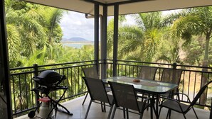 Ein Balkon mit Tisch und Stühlen, umgeben von üppiger Vegetation und Blick auf Strand und Berge.