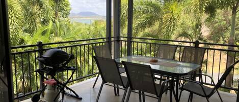 En balkon med et bord og stole, omgivet af frodig vegetation og udsigt til strand og bjerge.