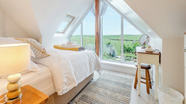 Une chambre à coucher avec un lit, une lampe de chevet et une vue sur un paysage verdoyant, visible à travers de grandes fenêtres.