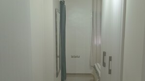 Ein schmaler Korridor mit weißen Wänden und Fliesenboden. Auf beiden Seiten befinden sich Türen, rechts ein Heizkörper und an der Decke eine Leuchte.