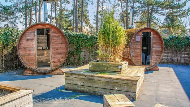 Due strutture a forma di botte in legno con porte, un piccolo tavolo in legno e una panchina in una zona boschiva.