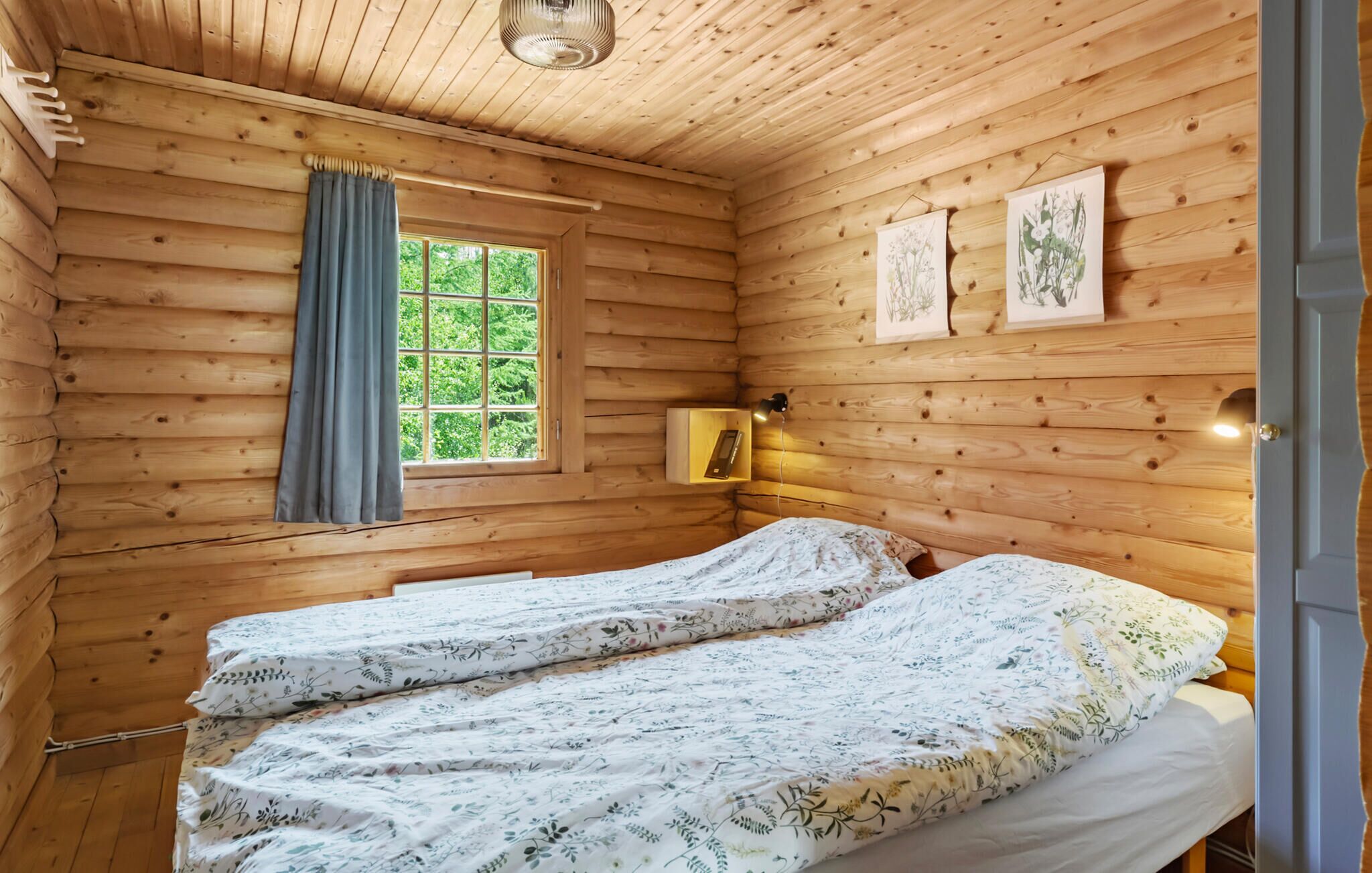 Ein Zimmer in einer Holzhütte mit zwei Betten, einem Fenster mit Vorhängen, einem Nachttisch mit Lampe und gerahmten Pflanzenbildern an der Wand.