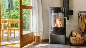 A wood-burning stove with a glass front, a wicker basket with firewood, and a wooden chair in a room with a glass door leading to an outdoor area. - Lovely home in Højslev with kitchen (Højslev)