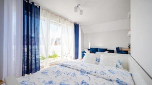 A bedroom with a large bed, a desk, and a chair. There is a window with curtains and a view of the outside. - 1 bedroom beach front apartment in Darlówko (Darlówko)