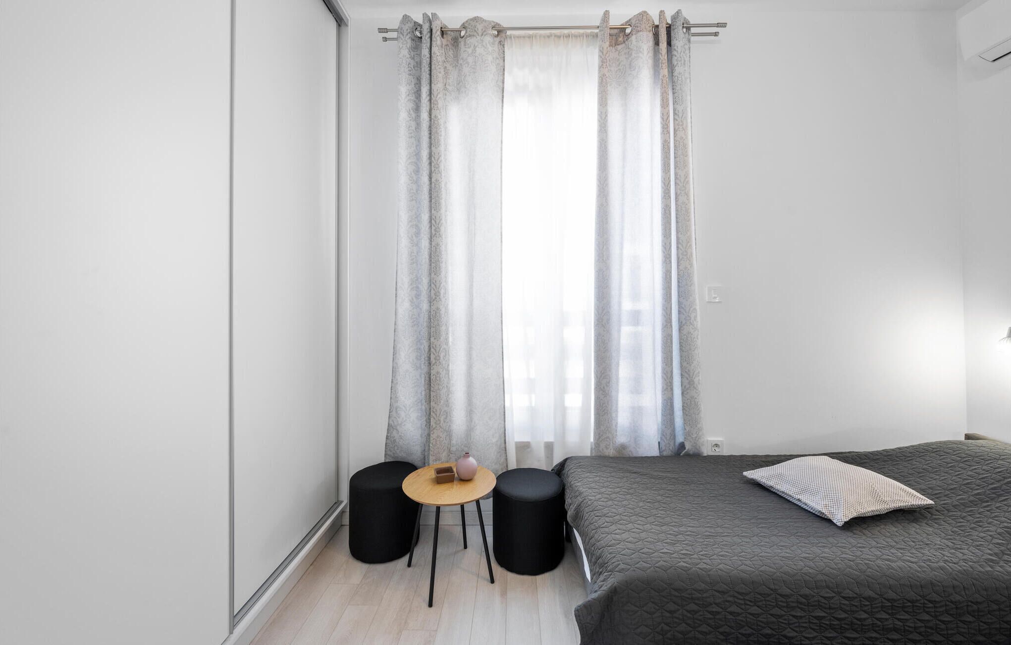 Ein Schlafzimmer mit einem Bett, zwei schwarzen Hocker, einem kleinen runden Tisch und einem Fenster mit durchsichtigen Vorhängen.