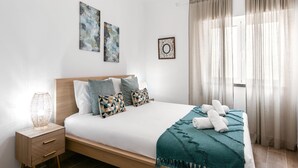 Una cama bien hecha con una manta y almohadas color turquesa, una mesita de noche con una lámpara y una obra de arte en la pared.