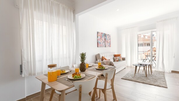 Un comedor moderno con una mesa de madera, sillas blancas y una preparación para el desayuno que incluye frutas y jugo.
