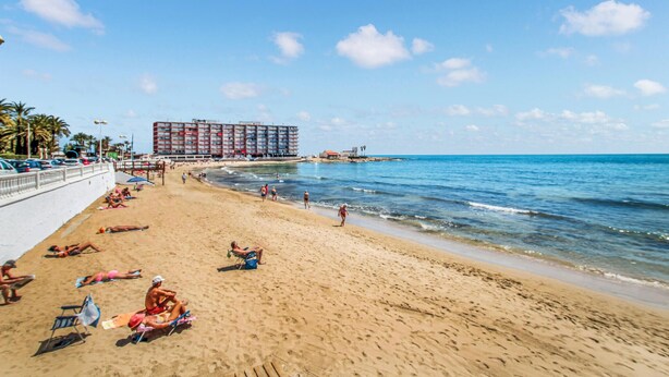 Una spiaggia con persone che prendono il sole e nuotano, un grande edificio alberghiero sullo sfondo e un cielo azzurro limpido.