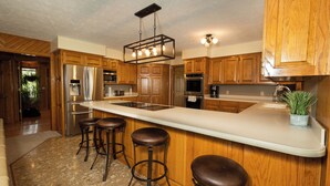 Una cocina con gabinetes de madera, una isla grande con taburetes, electrodomésticos de acero inoxidable y una lámpara colgante.