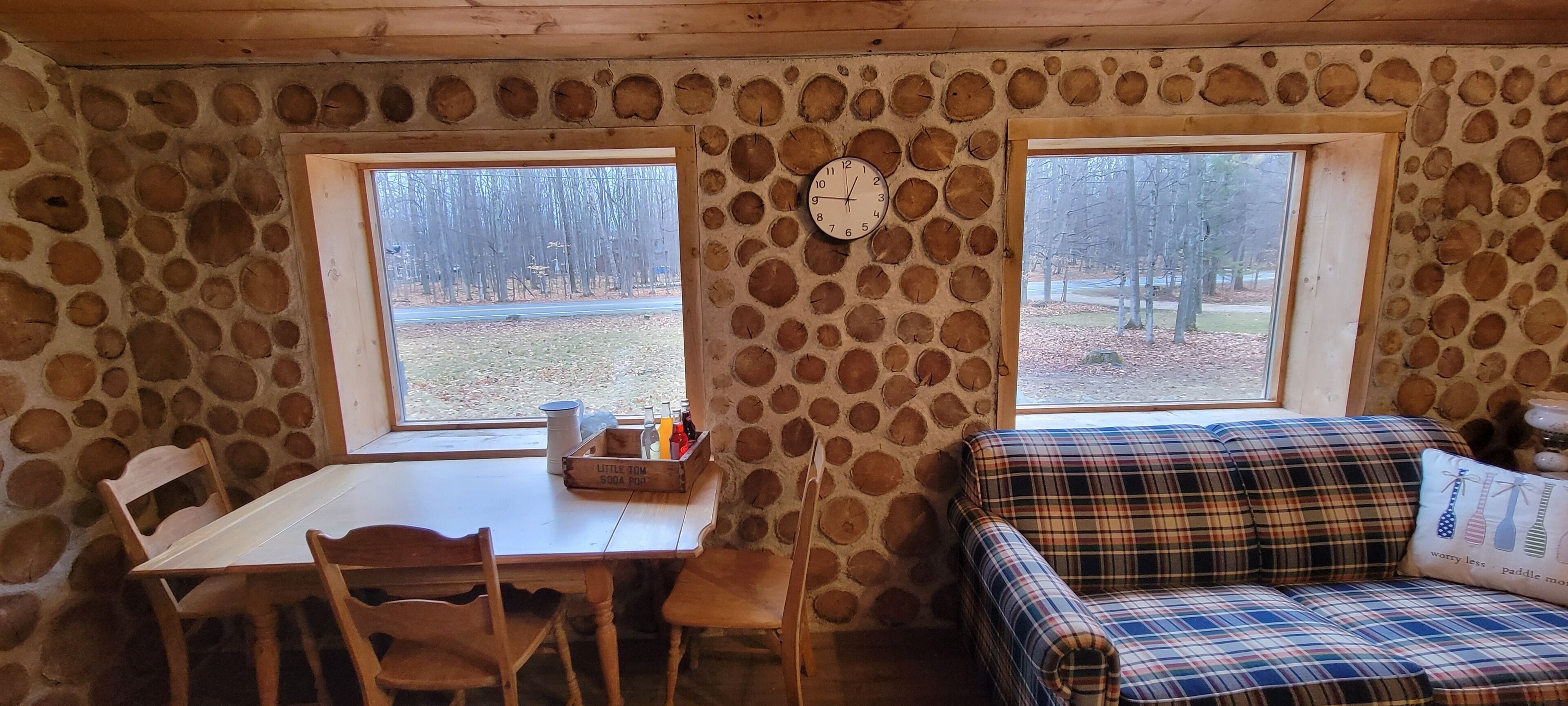 Interior d'una cabana rústica amb paret de pedra, mobles de fusta, taula de dinar, dues finestres i un rellotge.
