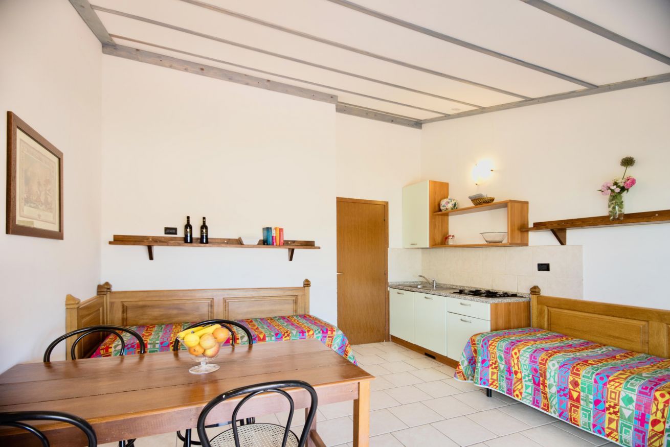 A compact room with a wooden dining table, two single beds, a kitchenette, and a small bathroom.