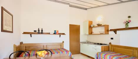 A compact room with a wooden dining table, two single beds, a kitchenette, and a small bathroom.