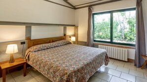 A bedroom with a bed, two bedside lamps, a window with curtains, and a radiator. - Two-room apartment for 4 people, 36 m2, consisting of:Living area with kitchenette (4 burners) and 2 single beds, double bedroom,bathroom with shower, covered terrace equipped with table, chairs and two deck chairs. (Guardistallo)