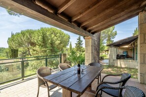 A wooden table with wicker chairs on a balcony overlooking a garden with trees and a stone building.