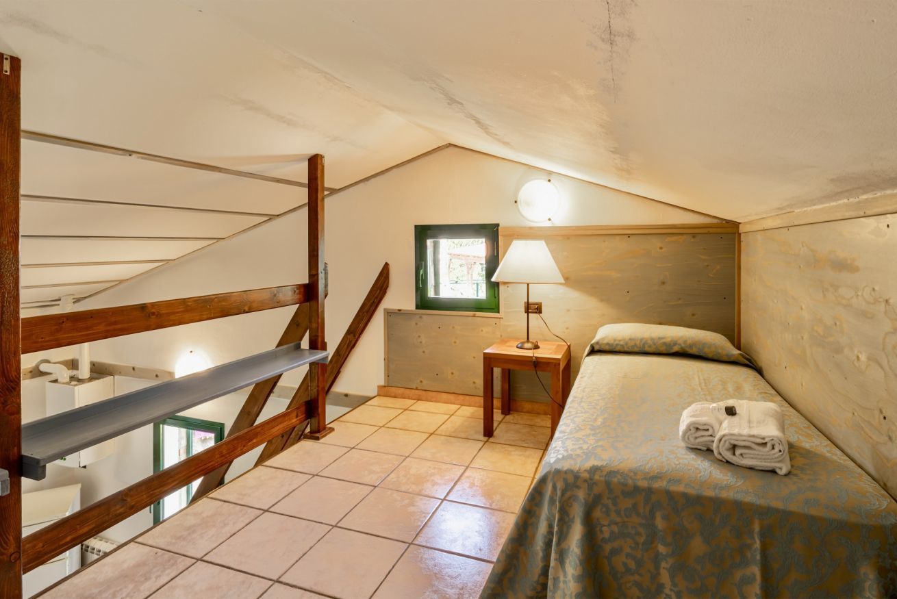 A small, cozy attic bedroom with a wooden staircase, a single bed with a patterned bedspread, a nightstand with a lamp, and a small window.