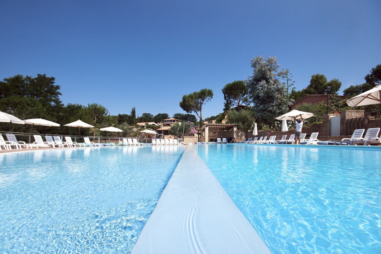 Una piscina azul cristalina con sillas de descanso blancas y sombrillas, rodeada de árboles y edificios.