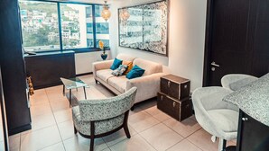 A modern living room with a sofa, two chairs, a small table, and a large window offering a city view. - Departamento en Puerto Santa Ana (Guayaquil)