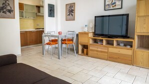 A living room with a sofa, a TV, a dining table with chairs, and a kitchen in the background. - M368 - Marcelli, new three-room apartment with garden (Marcelli)